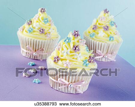 Stock Image - Thre Cupcakes Topped With Vanilla Butercream And Decorated With Blue, Pink And Purple Flowers, Pink Ribbon Tied Around Cake Cases, Purple And Blue Background. Fotosearch