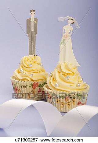 Stock Photo - Two Wedding Cupcakes With Piped Buttercream Topping Decorated With Pearls. Bride Figurine On One Cake And Groom On Other. Pale Blue/Lilac Background With White Ribbon. Fotosearch