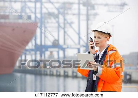 Stock Photo - Female engineer talking on walkie-talkie while using tablet PC in shipping yard. Fotosearch