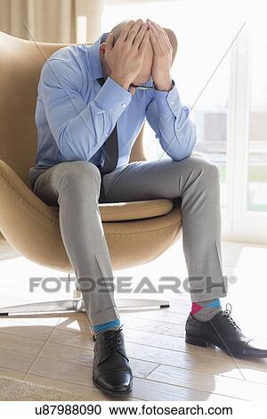 Stock Image - Full length of businessman with hands on head sitting on chair at home. Fotosearch