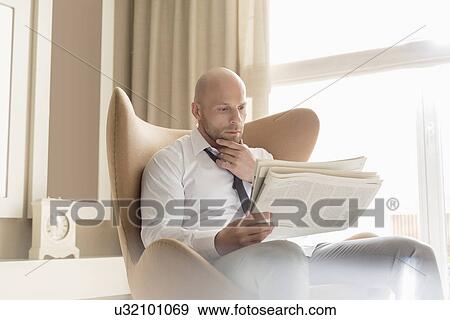 Stock Photo - Mid adult businessman reading newspaper on chair at home. Fotosearch