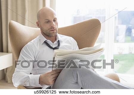 Stock Photography - Mid adult businessman reading newspaper on wing back chair at home. Fotosearch