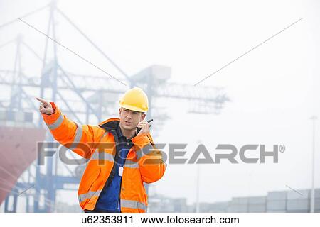 Stock Image - Mid adult man pointing while using walkie-talkie in shipping yard. Fotosearch