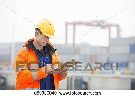 Stock Image - Mid adult man writing on clipboard in shipping yard. Fotosearch
