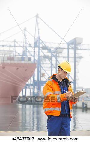 Stock Photo - Mid adult man writing on clipboard in shipping yard. Fotosearch