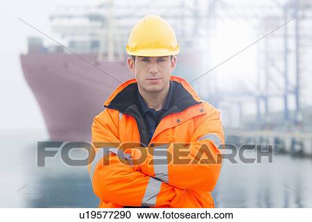 Stock Image - Portrait of confident mid adult man wearing protective clothing in shipping yard. Fotosearch