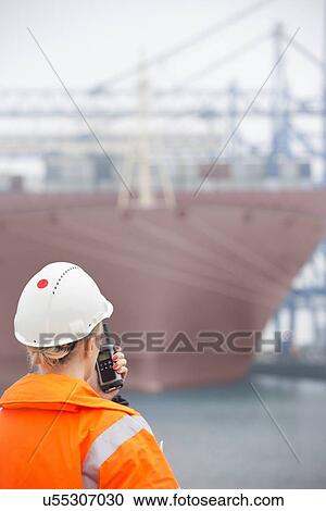Stock Image - Rear view of female engineer using walkie-talkie in shipping yard. Fotosearch