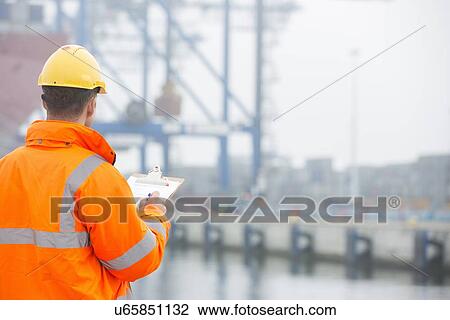 Stock Image - Rear view of mid adult man writing on clipboard in shipping yard. Fotosearch