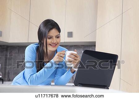 Stock Photo - Beautiful woman with coffee mug looking at laptop in kitchen. Fotosearch