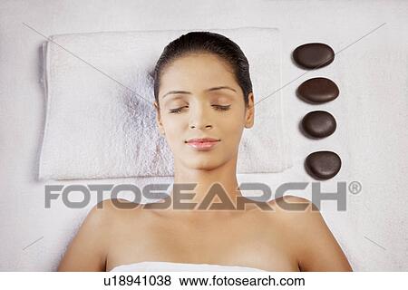 Stock Photo - Close-up of young woman lying on massage table with pebble stones on side . Fotosearch