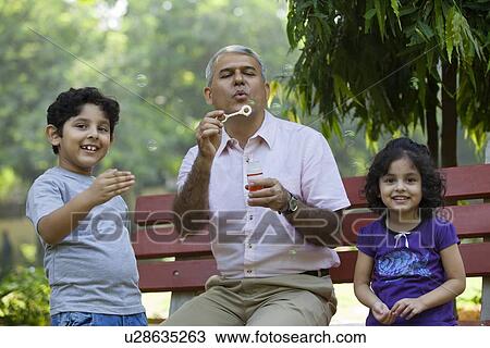 Stock Image - Grandfather blowing bubbles with grandchildren in park. Fotosearch