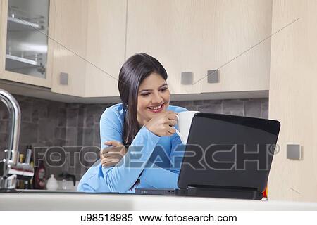 Happy Indian woman with coffee mug looking at laptop in kitchen View Large Photo Image Stock Photography - Happy Indian woman with coffee mug looking at laptop in kitchen. Fotosearch