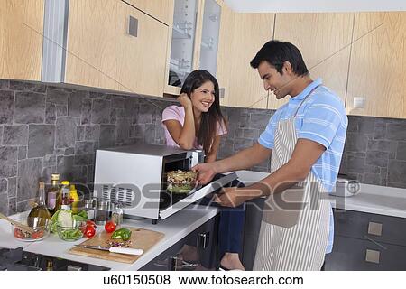 Stock Photo - Happy woman looking at man preparing food in kitchen. Fotosearch