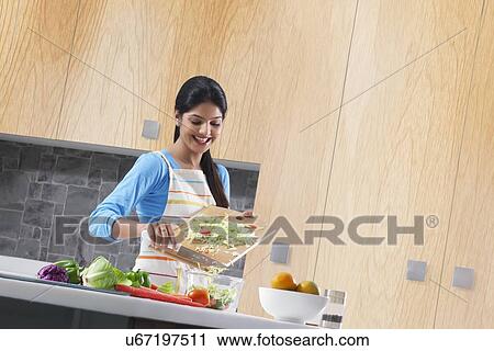 Stock Image - Happy young woman preparing vegetable salad in kitchen. Fotosearch