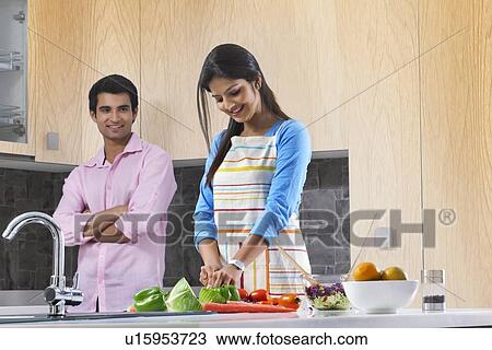 Stock Image - Man looking at woman cutting vegetables in kitchen. Fotosearch