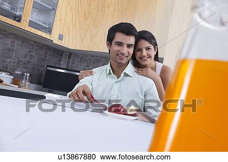 Stock Image - Portrait of affectionate couple with apple and catalogues in kitchen. Fotosearch