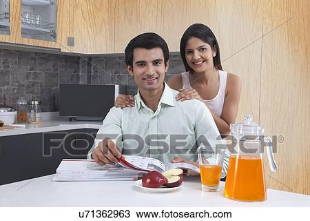 Stock Image - Portrait of Indian couple with catalogues at kitchen island. Fotosearch