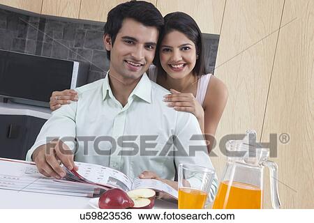 Stock Photograph - Portrait of loving couple with catalogues at kitchen island. Fotosearch