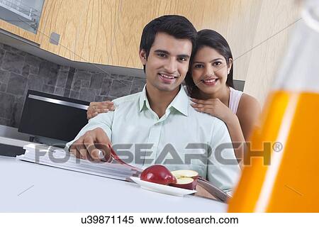 Stock Photography - Portrait of smiling couple with catalogues and apple at kitchen island. Fotosearch