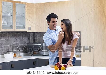 Stock Image - Smiling man feeding fresh apple to woman in kitchen. Fotosearch
