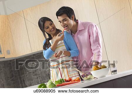 Stock Photography - Smiling woman feeding carrot to man in kitchen. Fotosearch