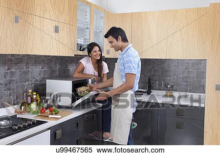 Stock Photography - Smiling woman looking at man preparing meal in kitchen. Fotosearch