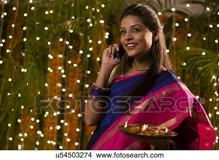 Woman talking on a mobile phone while holding a tray of diyas View Large Photo Image Picture - Woman talking on a mobile phone while holding a tray of diyas . Fotosearch