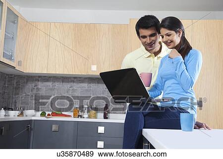 Stock Photo - Young Indian couple using laptop in kitchen. Fotosearch