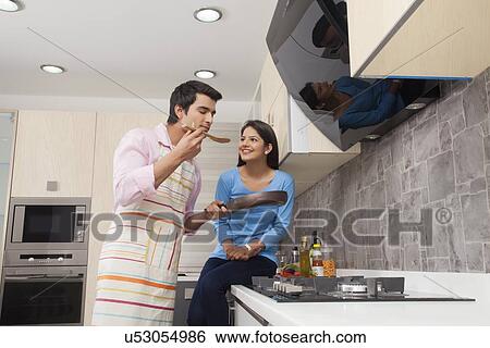 Stock Photograph - Young man smelling food while woman sitting at kitchen counter. Fotosearch