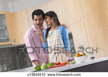 Stock Image - Young woman feeding carrot to man in kitchen. Fotosearch