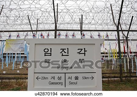Picture - Barbed wire fence separates South from North Korea - South Korean flags attached to fence - Asia - SIGN POINTS TO GAESEONG AND SEOUL. Fotosearch