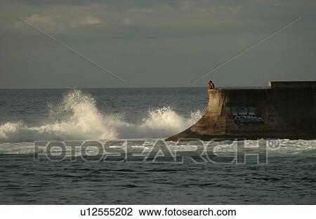 Stock Image - A man sitting on a reef watching the surf, Papeete, Tahiti. Fotosearch