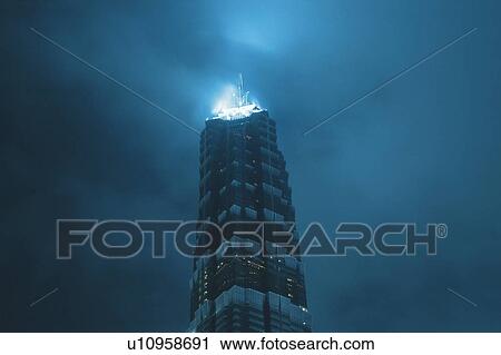 Stock Image -  the Jin Mao Tower Under a Cloudy Sky, the Top Shining in a Blue Color, Low Angle View, Shanghai, China. Fotosearch