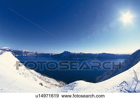 Stock Photo -  the Lake Mashu, Surrounded By Mountainchains Covered In Snow, the Sun Shining in Addition, Hokkaido Prefecture, Japan, Lens Flare. Fotosearch