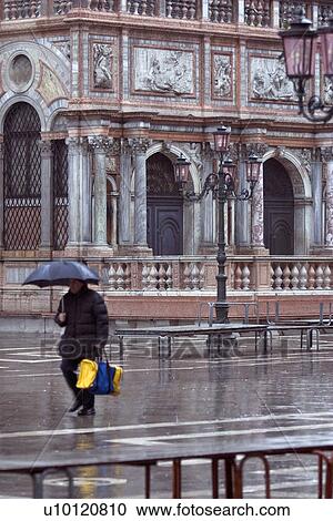Stock Image - Man walking in rain with sports-bag and umbrella in St Marks Piazza. Fotosearch