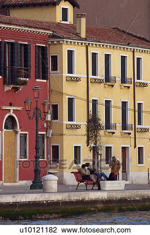 Stock Image - yellow and red buildings with ornamental lamp post, people seated on bench at edge of canal. Fotosearch