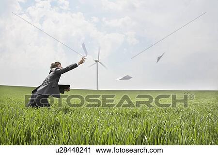 Stock Image - Businessman chasing papers on wind farm. Fotosearch