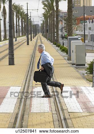 Stock Image - Senior businessman running across pedestrian crossing on tramway. Fotosearch
