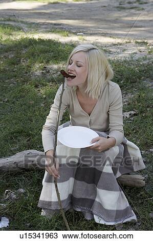 Stock Image - Woman at campsite eating a hotdog off a stick and smiling.. Fotosearch