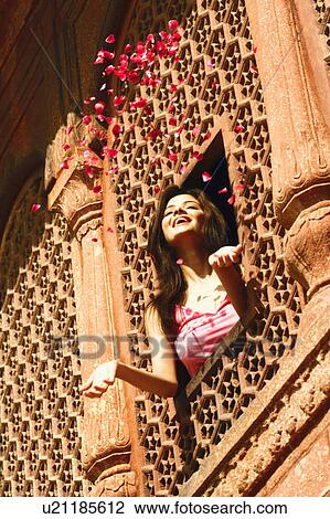 Stock Image - Low angle view of a young woman throwing rose petal from a window. Fotosearch