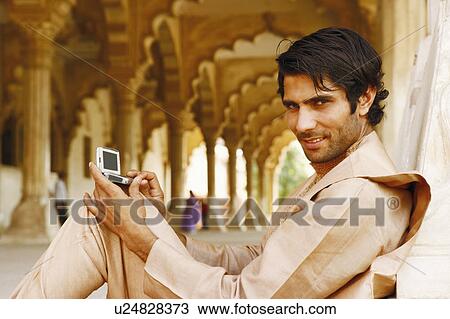 Stock Image - Portrait of a young man holding a hand-held device, Agra Fort, Agra, Uttar Pradesh, India. Fotosearch