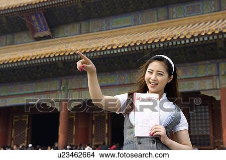 Picture - Smiling young woman pointing, standing in front of a palace building in the Forbidden City, Beijing, China. Fotosearch