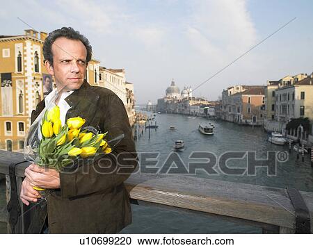 Stock Image - Italy, Venice, man waiting on bridge holding flowers. Fotosearch