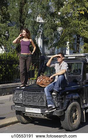 Stock Image - Woman standing on hood of SUV on cellular phone with man sitting on hood.. Fotosearch