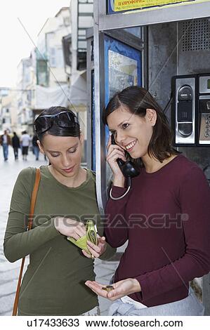 Stock Image - Two young girls on public phone one with wallet in hand the other on the phone talking city in background.. Fotosearch