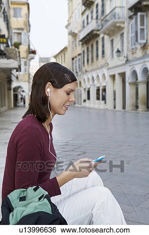 Young girl sitting on steps in street earphones in ears MP3 in hand smiling old city in background. View Large Photo Image Stock Photograph - Young girl sitting on steps in street earphones in ears MP3 in hand smiling old city in background.. Fotosearch