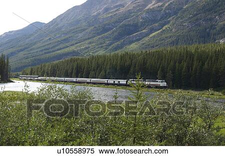 Stock Photography - A train passing by a lake and mountains, Rocky Mountains, Canada. Fotosearch