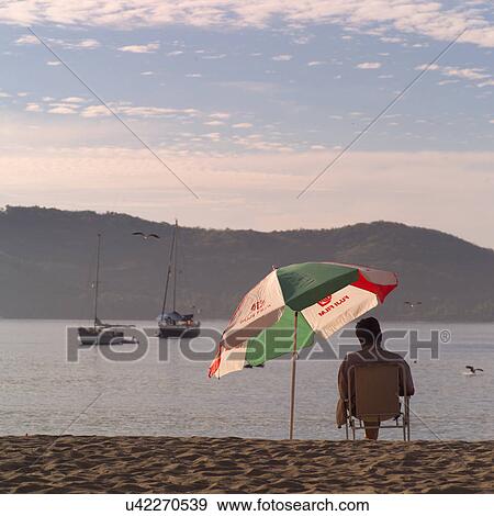 Stock Photo - Person sitting under an umbrella on a beach. Fotosearch
