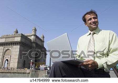 Stock Photo - Low angle view of a businessman holding a laptop and smiling with a monument in the background, Gateway of India, Mumbai, Maharashtra, India. Fotosearch