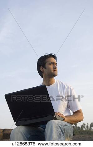 Stock Photograph - Low angle view of a young man holding a laptop on his lap and sitting at a coast, Madh Island, Mumbai, Maharashtra, India. Fotosearch